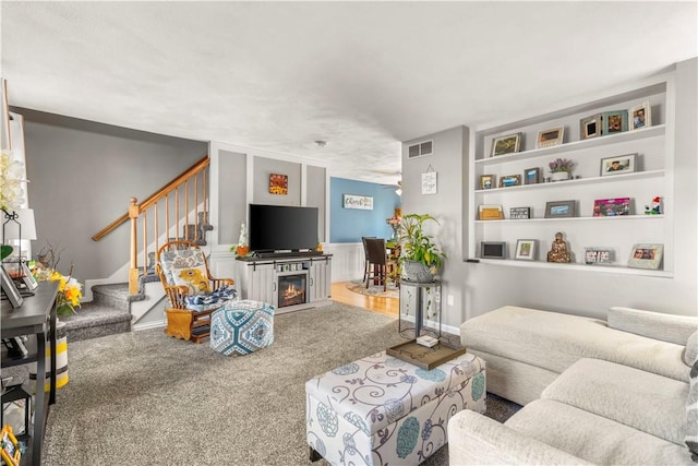 carpeted living area featuring built in shelves, visible vents, a lit fireplace, baseboards, and stairs