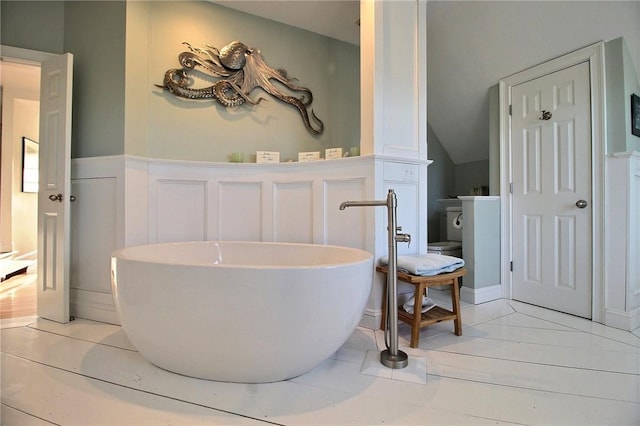 bathroom with a soaking tub, vaulted ceiling, a decorative wall, and wainscoting