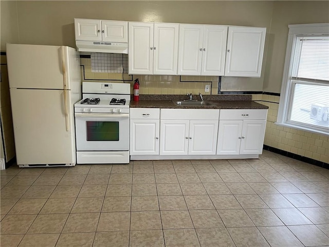 kitchen featuring white appliances, dark countertops, under cabinet range hood, a sink, and light tile patterned flooring