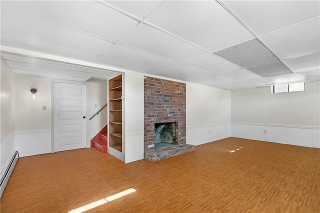 finished basement with a drop ceiling, baseboard heating, wainscoting, and a brick fireplace