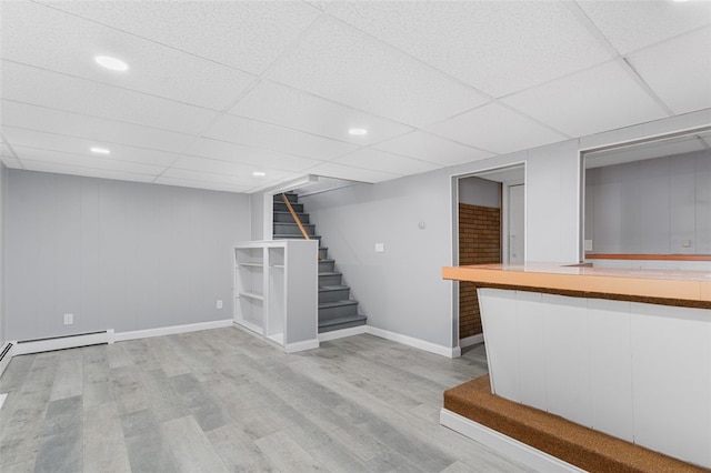 finished basement with baseboards, light wood-style flooring, stairs, a paneled ceiling, and a baseboard heating unit