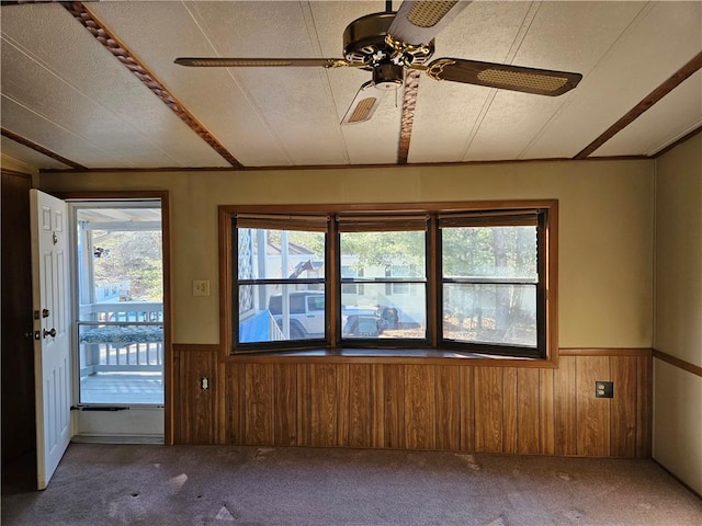 carpeted empty room with a textured ceiling, wooden walls, a ceiling fan, and wainscoting