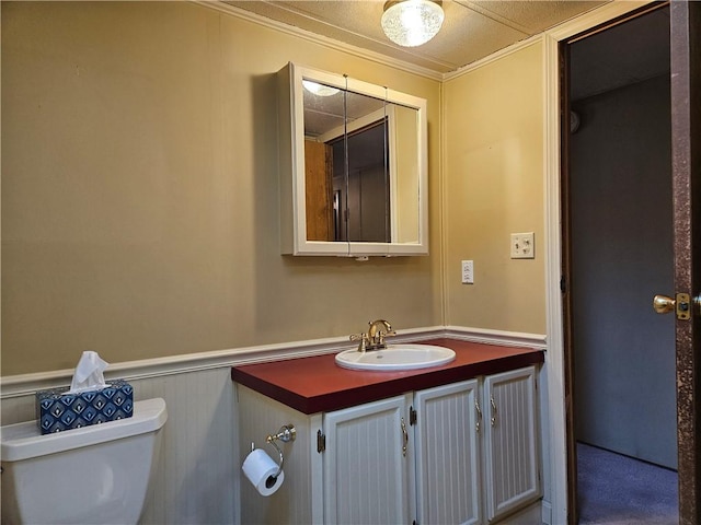 half bathroom featuring toilet, a wainscoted wall, crown molding, vanity, and a decorative wall