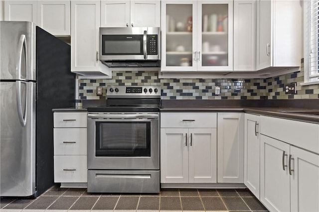 kitchen with dark countertops, glass insert cabinets, stainless steel appliances, white cabinetry, and backsplash