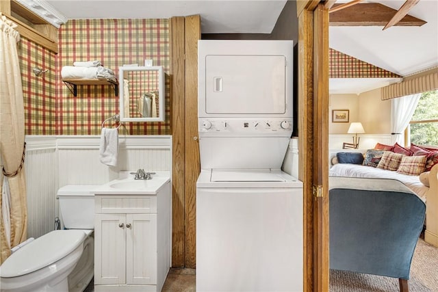 washroom with wallpapered walls, laundry area, stacked washer and clothes dryer, a wainscoted wall, and a sink