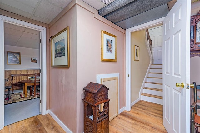 corridor featuring a drop ceiling, wooden walls, baseboards, stairway, and light wood finished floors