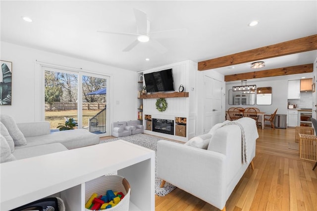 living area featuring light wood-style floors, a glass covered fireplace, beamed ceiling, and ceiling fan with notable chandelier