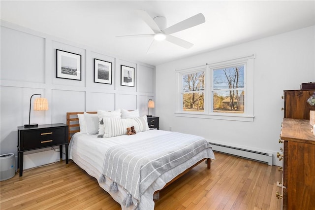 bedroom featuring light wood-style flooring, baseboard heating, a ceiling fan, and a decorative wall