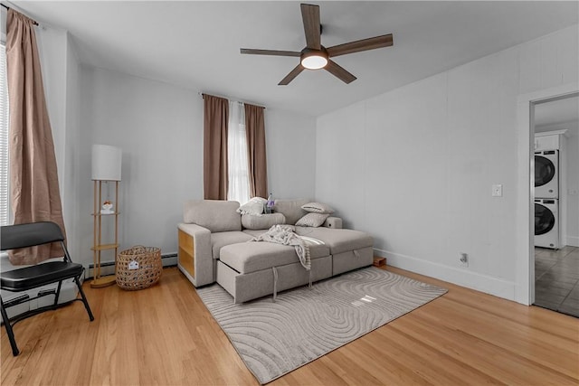 living area featuring a baseboard heating unit, wood finished floors, a ceiling fan, baseboards, and stacked washing maching and dryer