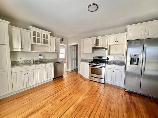 kitchen with stone counters, white cabinetry, light wood-style floors, appliances with stainless steel finishes, and glass insert cabinets