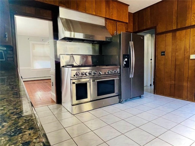 kitchen featuring brown cabinets, stainless steel appliances, a baseboard radiator, wood walls, and wall chimney range hood