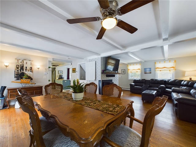 dining area featuring a baseboard heating unit, wood finished floors, a ceiling fan, a brick fireplace, and beam ceiling