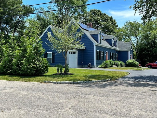 cape cod house featuring an attached garage, a front lawn, roof mounted solar panels, a chimney, and driveway