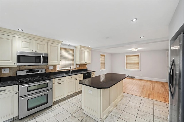 kitchen featuring a sink, appliances with stainless steel finishes, cream cabinetry, decorative backsplash, and dark countertops