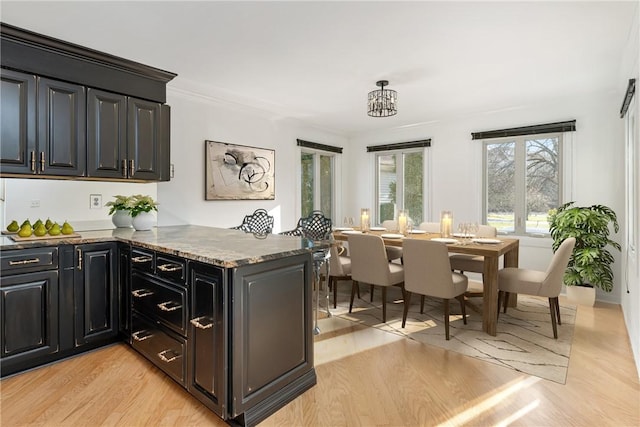 kitchen with ornamental molding, a peninsula, dark cabinetry, light wood-type flooring, and stone counters