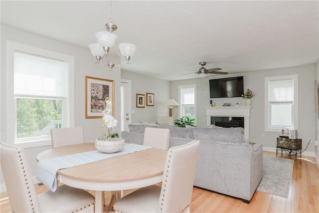 dining area featuring light wood-style flooring, a fireplace, ceiling fan with notable chandelier, and baseboards