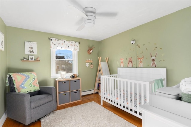 bedroom featuring a crib, wood finished floors, a ceiling fan, and a baseboard radiator