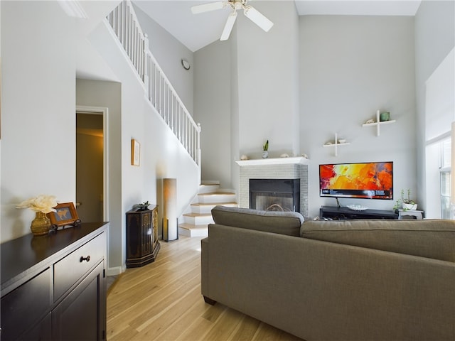 living room with stairway, a ceiling fan, a high ceiling, light wood-style floors, and a brick fireplace