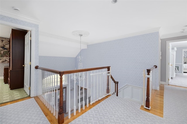 hall with wood finished floors, wallpapered walls, ornamental molding, wainscoting, and an upstairs landing