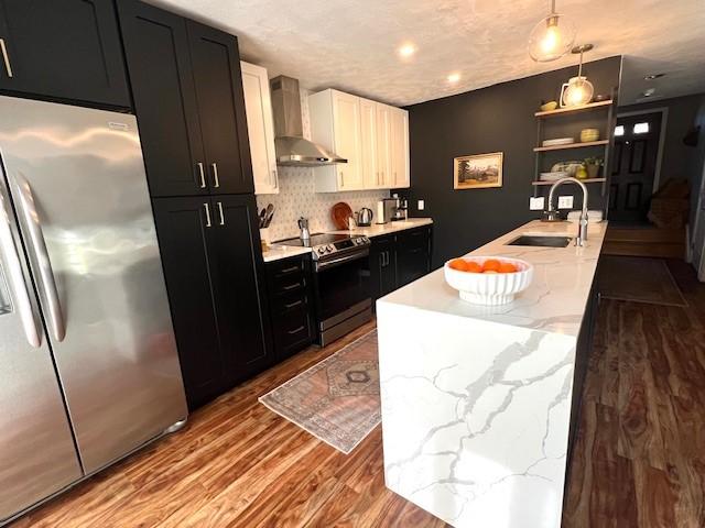 kitchen featuring a sink, open shelves, stainless steel appliances, wall chimney exhaust hood, and dark cabinets