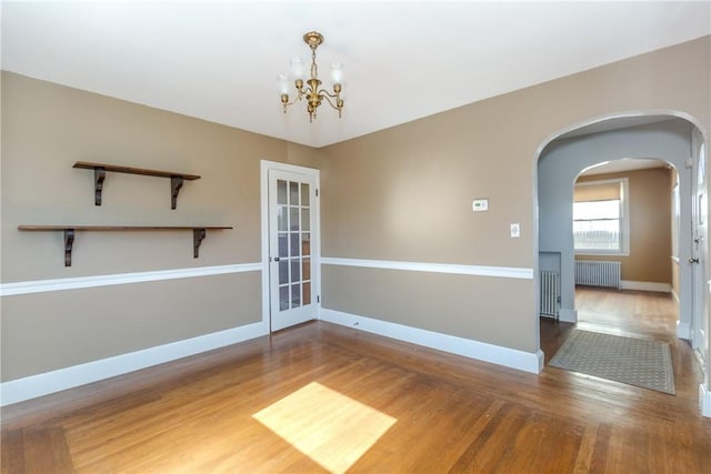 empty room with a notable chandelier, wood finished floors, radiator heating unit, arched walkways, and baseboards