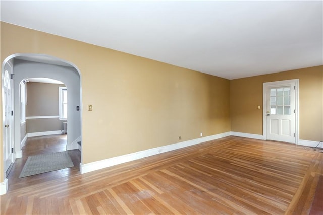 empty room featuring arched walkways, a wealth of natural light, light wood-style floors, and baseboards