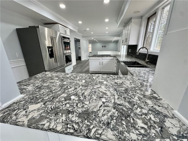 kitchen with a peninsula, dark stone counters, a sink, appliances with stainless steel finishes, and crown molding