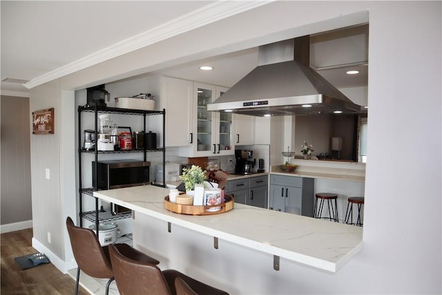 kitchen with a kitchen breakfast bar, island range hood, crown molding, glass insert cabinets, and light stone countertops