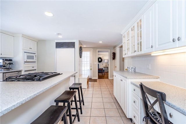 kitchen with light tile patterned floors, white microwave, a breakfast bar, glass insert cabinets, and backsplash