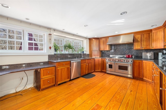 kitchen with appliances with stainless steel finishes, wall chimney range hood, brown cabinetry, and recessed lighting