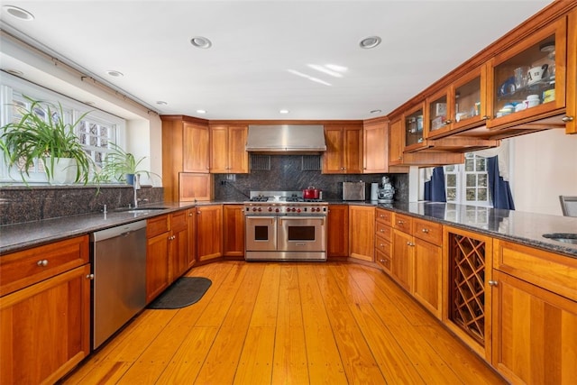 kitchen featuring appliances with stainless steel finishes, wall chimney range hood, a sink, brown cabinetry, and tasteful backsplash