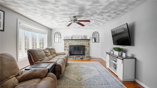 living room featuring baseboards, a stone fireplace, wood finished floors, a textured ceiling, and a ceiling fan