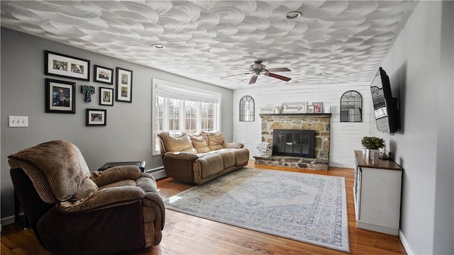 living area featuring a baseboard heating unit, wood finished floors, a stone fireplace, baseboards, and ceiling fan