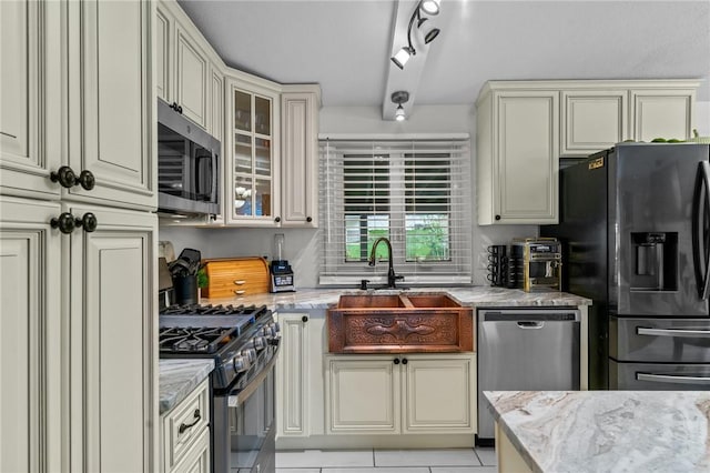 kitchen with cream cabinetry, a sink, light stone counters, stainless steel appliances, and glass insert cabinets