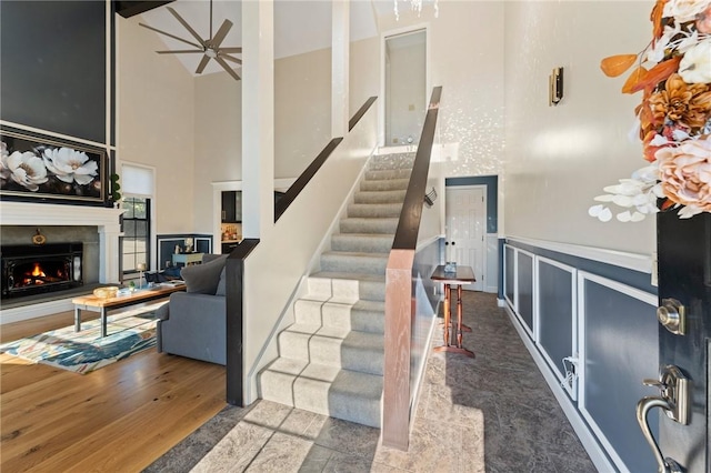 staircase featuring a wainscoted wall, a warm lit fireplace, wood finished floors, a high ceiling, and ceiling fan