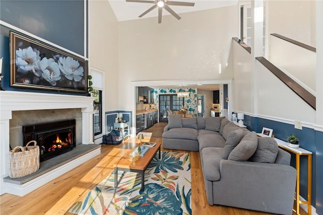 living area featuring a high ceiling, wood finished floors, a warm lit fireplace, and ceiling fan