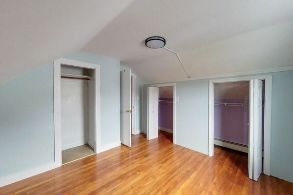 unfurnished bedroom with vaulted ceiling, light wood-style flooring, multiple closets, and baseboard heating