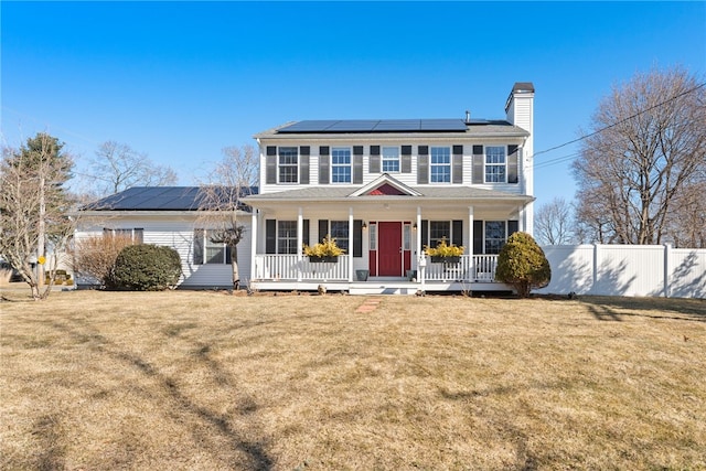 colonial inspired home featuring roof mounted solar panels, covered porch, a front lawn, and fence