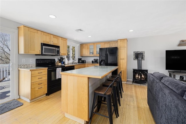 kitchen featuring visible vents, black appliances, light brown cabinetry, open floor plan, and a wood stove