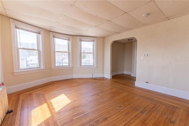 empty room with radiator, baseboards, light wood-style flooring, arched walkways, and a paneled ceiling