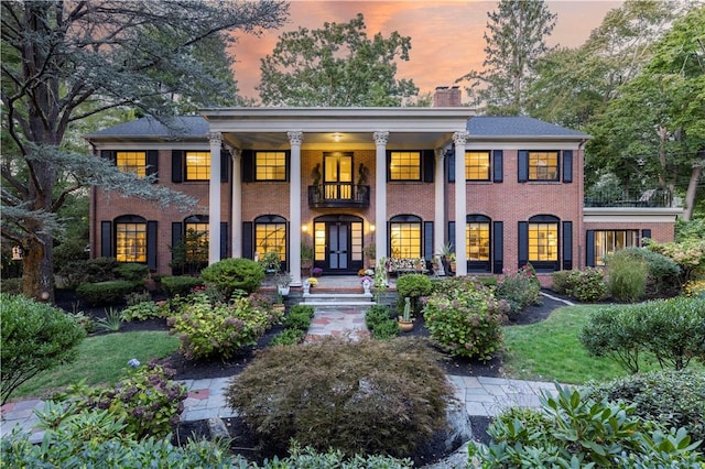 greek revival inspired property featuring brick siding, covered porch, a chimney, and a balcony