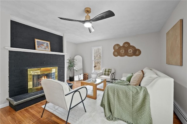 living area featuring a ceiling fan, a baseboard heating unit, wood finished floors, baseboards, and a brick fireplace