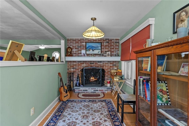 interior space featuring a brick fireplace, wood finished floors, baseboards, and ceiling fan