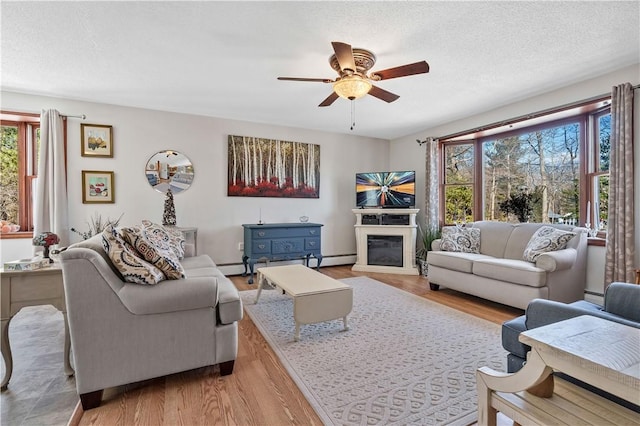 living room with light wood finished floors, ceiling fan, a textured ceiling, a glass covered fireplace, and baseboard heating