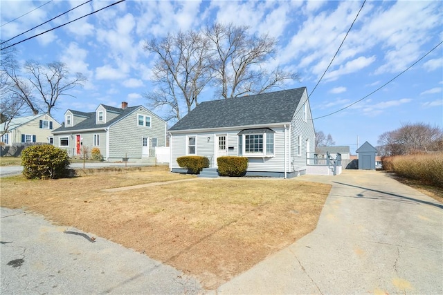 cape cod home with a front lawn, concrete driveway, an outbuilding, and roof with shingles