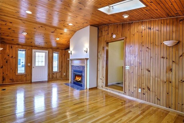 unfurnished living room with a premium fireplace, vaulted ceiling with skylight, light wood-style flooring, wood ceiling, and wood walls