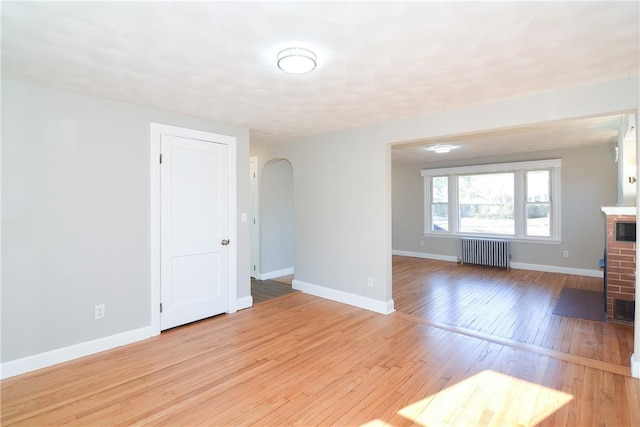 spare room featuring arched walkways, light wood-style floors, radiator, baseboards, and a brick fireplace