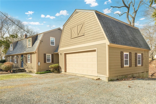 view of front of home with gravel driveway, a gambrel roof, roof with shingles, a garage, and an outbuilding