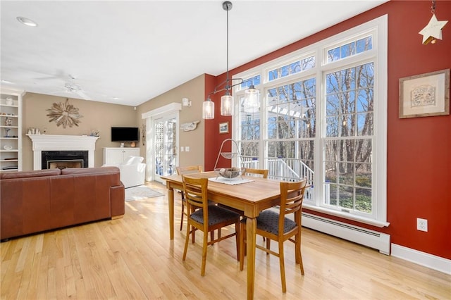 dining room with light wood-type flooring, a ceiling fan, a fireplace, a baseboard radiator, and baseboards