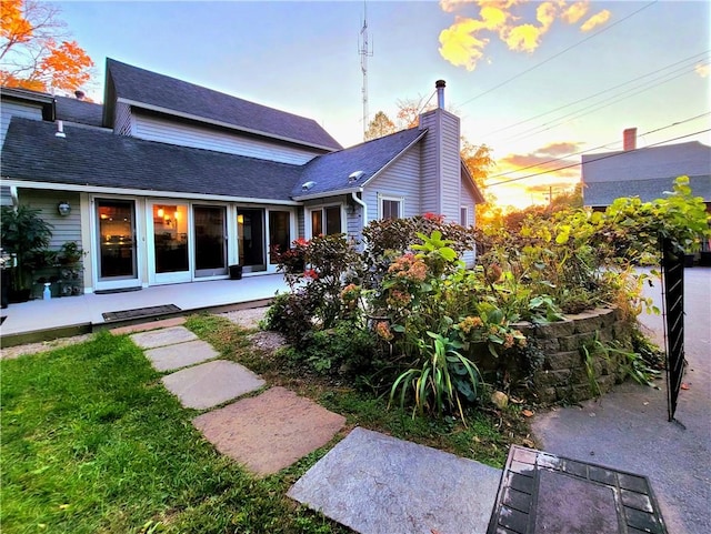 back of house at dusk featuring a patio, roof with shingles, and a chimney